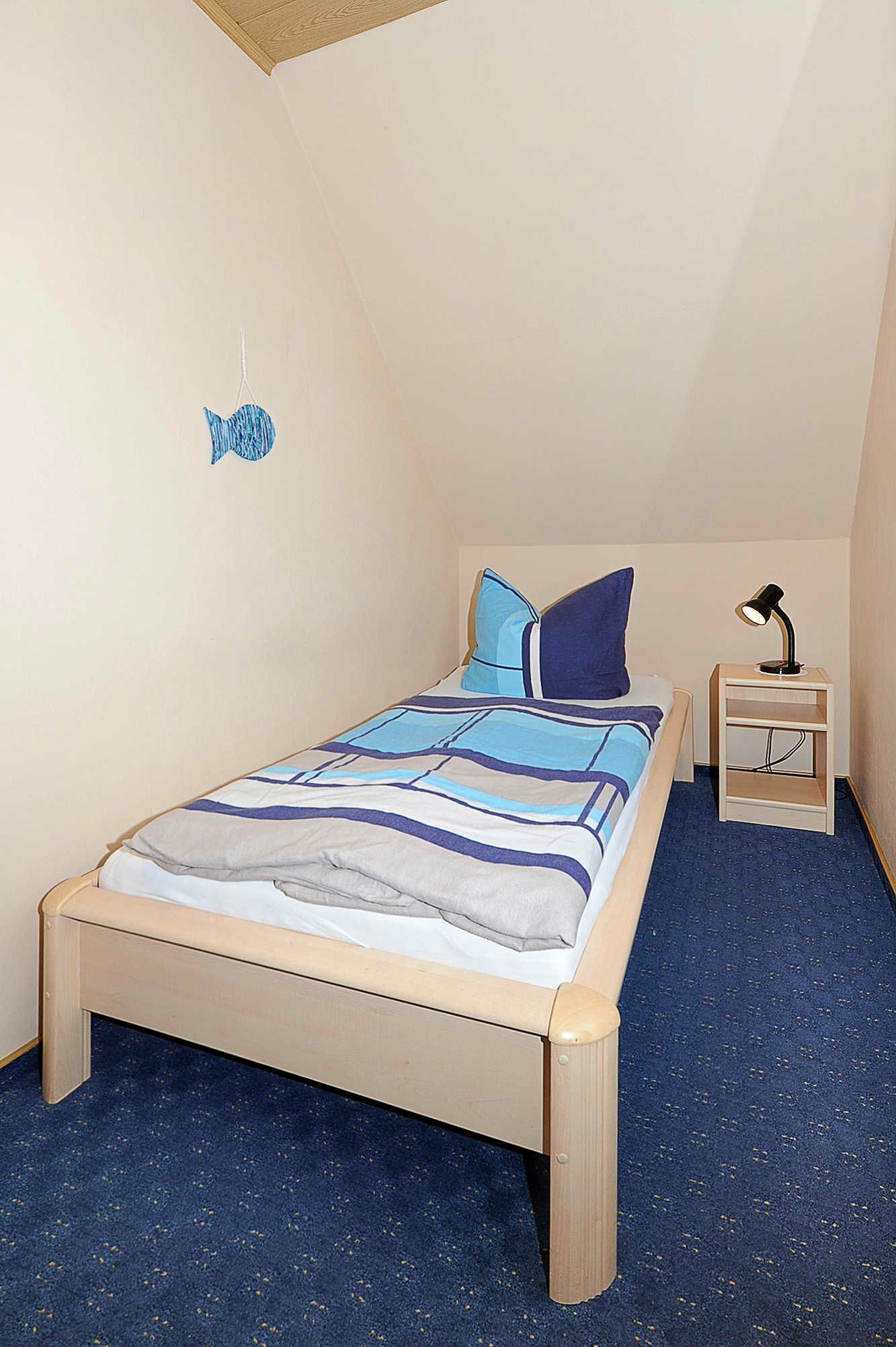 A single bed with a blue and white striped bedspread, a small bedside table with a lamp, and a wall decoration of two fish.