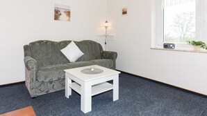 A room with a green patterned sofa, a white coffee table, a lamp, and a window with a view of trees. - Ferienwohnung 2 im Ferienhaus Trautes Heim - Ferienwohnung 2 (Esens)