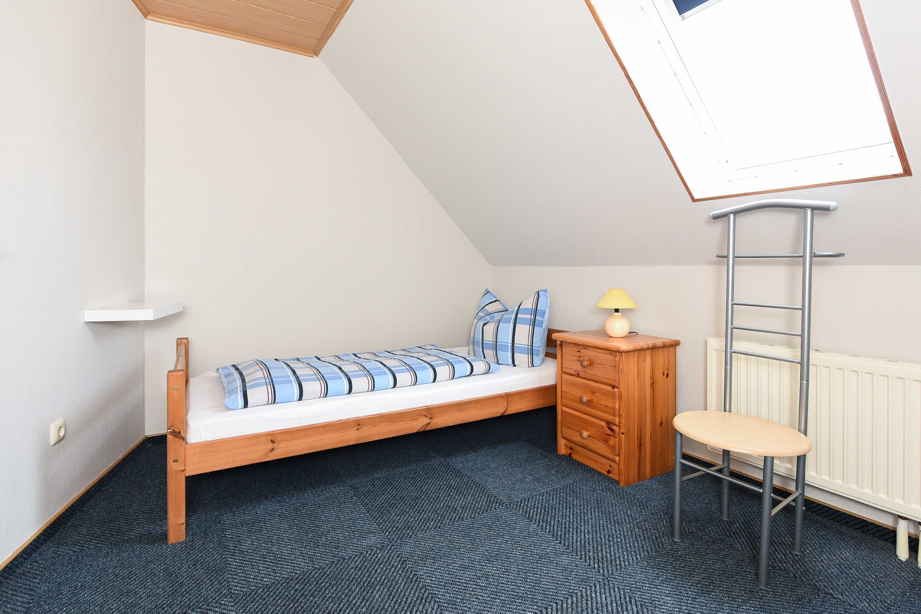 A small, single-bed room with a sloped ceiling, a wooden dresser, a small table with a lamp, and a radiator.