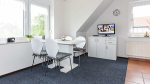 A modern kitchen with white cabinetry, a dining table with chairs, a television, and a window with a view of the outside. - Ferienwohnung 2 im Ferienhaus Trautes Heim - Ferienwohnung 2 (Esens)