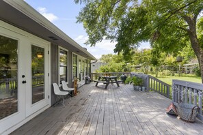 Una terraza de madera con una mesa y sillas de picnic, rodeada por un edificio blanco con ventanas grandes y una barandilla gris.