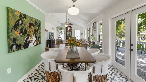Un coin repas avec une table en bois, des chaises blanches et un vase rempli de tournesols. On y trouve également un ventilateur de plafond, un tableau accroché au mur et une vue sur l’extérieur grâce à de grandes fenêtres.