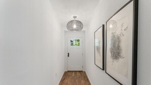 Un couloir doté d’un plancher en bois, de murs blancs, d’une porte blanche avec une fenêtre et de deux œuvres d’art encadrées.