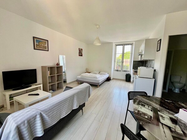 Una habitación pequeña con cama, televisión, mesita, estantería y ventana.