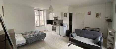 A compact living space with a bed, sofa, small table, kitchenette, and a door leading to another room.