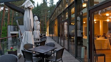 Area esterna con zona pranzo, tavoli e sedie, coperta da ombrelloni e con una parete di vetro che si affaccia su un ristorante interno.
