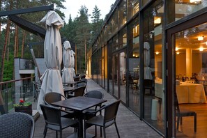 Un espacio de comedor al aire libre con mesas y sillas, cubierto por sombrillas, y una pared de vidrio que da a un restaurante en el interior.
