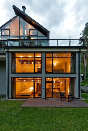Una casa moderna de dos pisos con grandes ventanales, un interior visible con cama y zona de estar, y una terraza al frente.