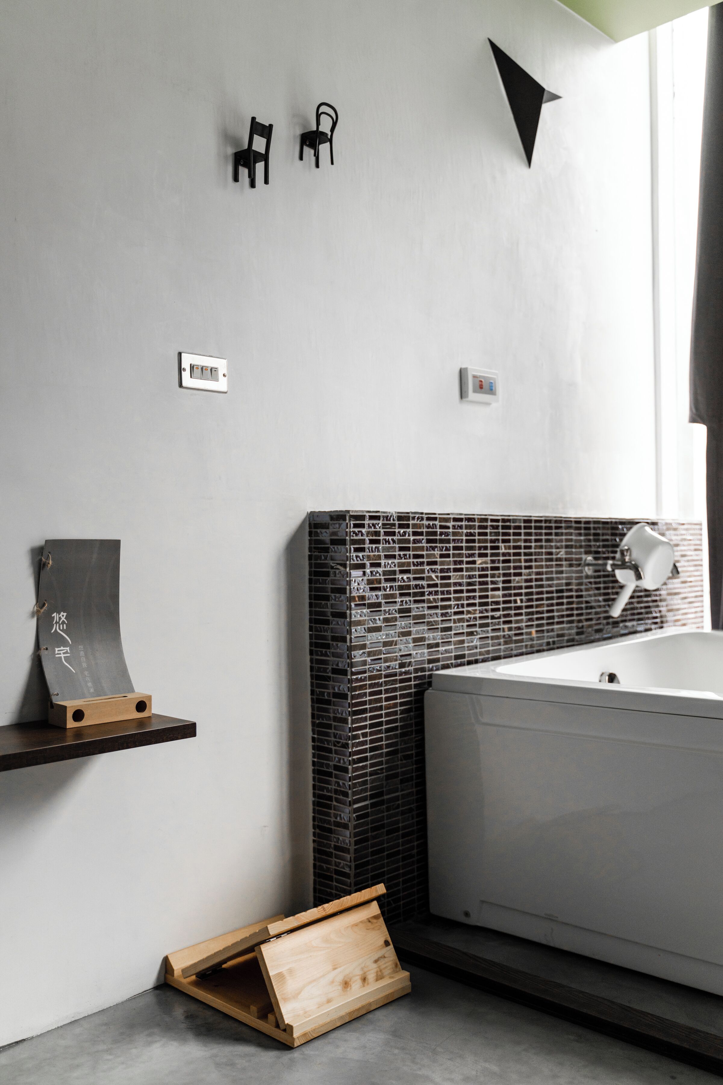 A modern bathroom with a bathtub, a wooden shelf with a sculpture, and a black chair mounted on the wall.