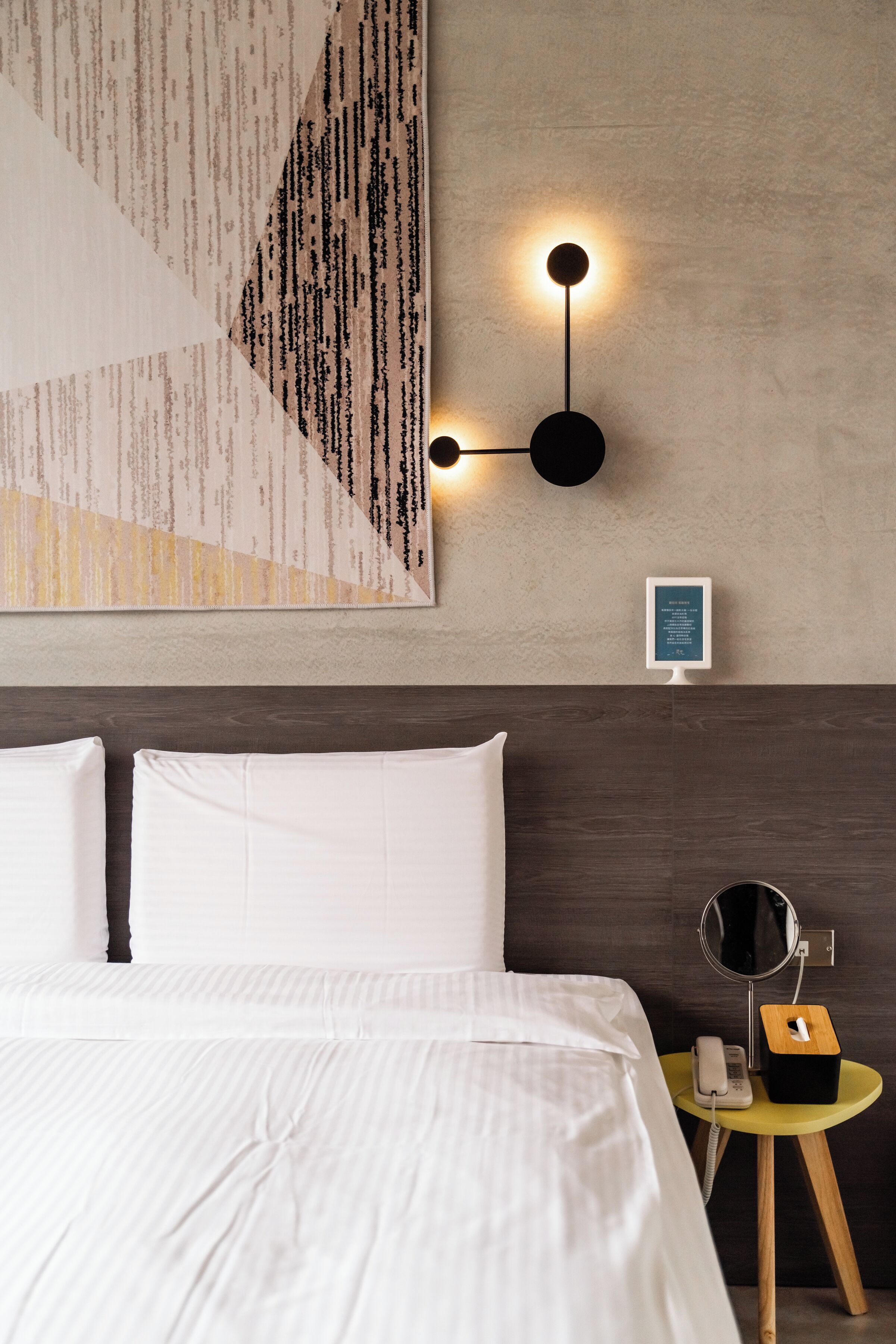 Un lit bien fait, avec du linge de lit blanc, un luminaire fixé au mur, une petite table de chevet avec un téléphone et une boîte, et un grand tableau abstrait accroché au mur.