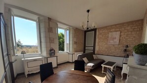 A cozy room with a round wooden table, a sofa, and a small table. There is a window with a view of greenery outside. - Quiet cottage with garden and WIFI in La Roche-Posay (La Roche-Posay)