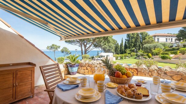 A table set for breakfast with a view of the sea, a stone wall, and a wooden chair.