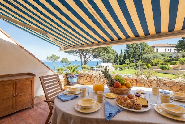 A table set for breakfast with a view of the sea, a stone wall, and a wooden chair.