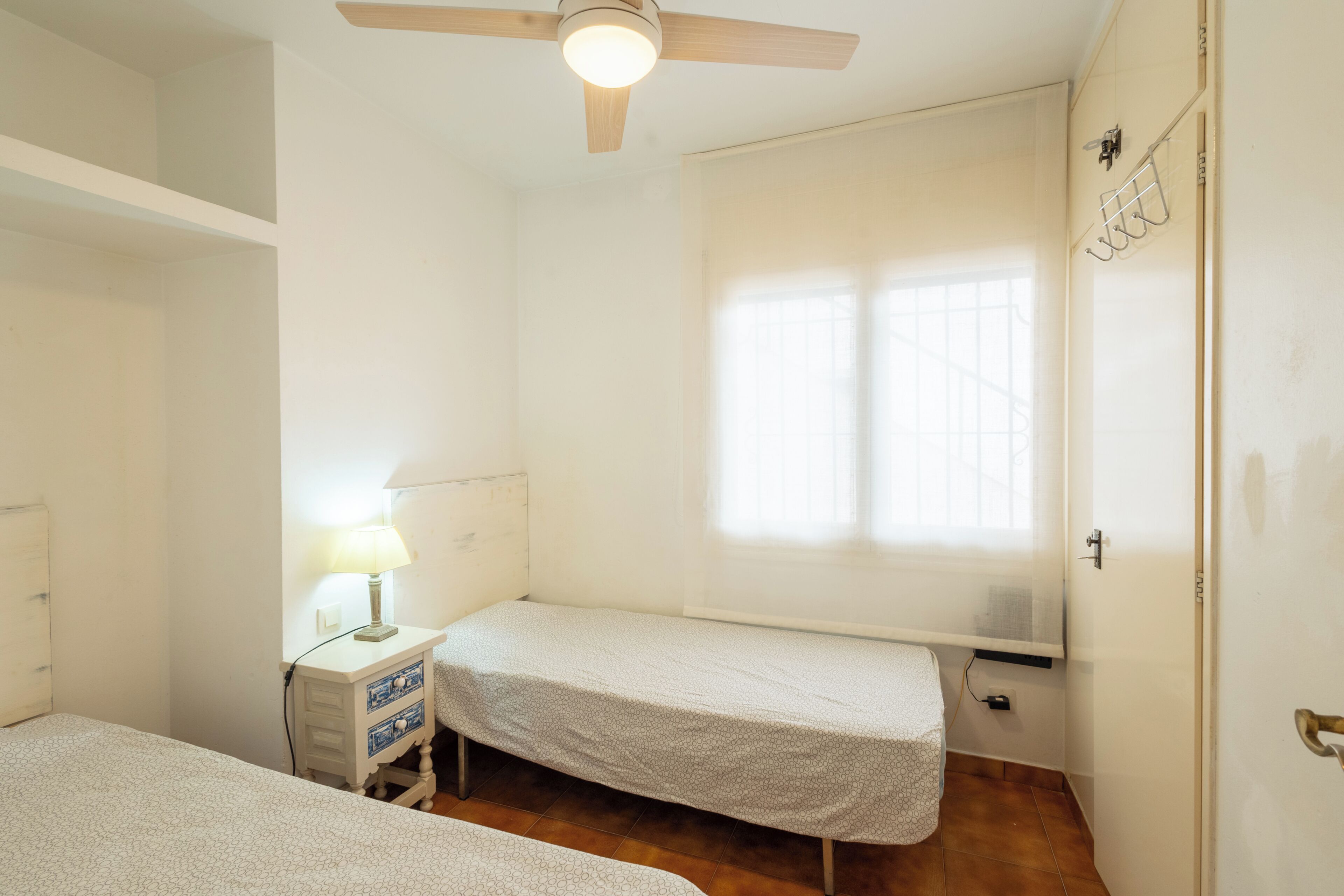 Un petit logement composé d’une seule pièce, comprenant un lit, une table de chevet, une lampe et un ventilateur de plafond.
