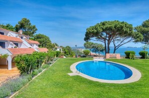 A swimming pool surrounded by a well-maintained lawn, with a building featuring red-tiled roofs in the background.