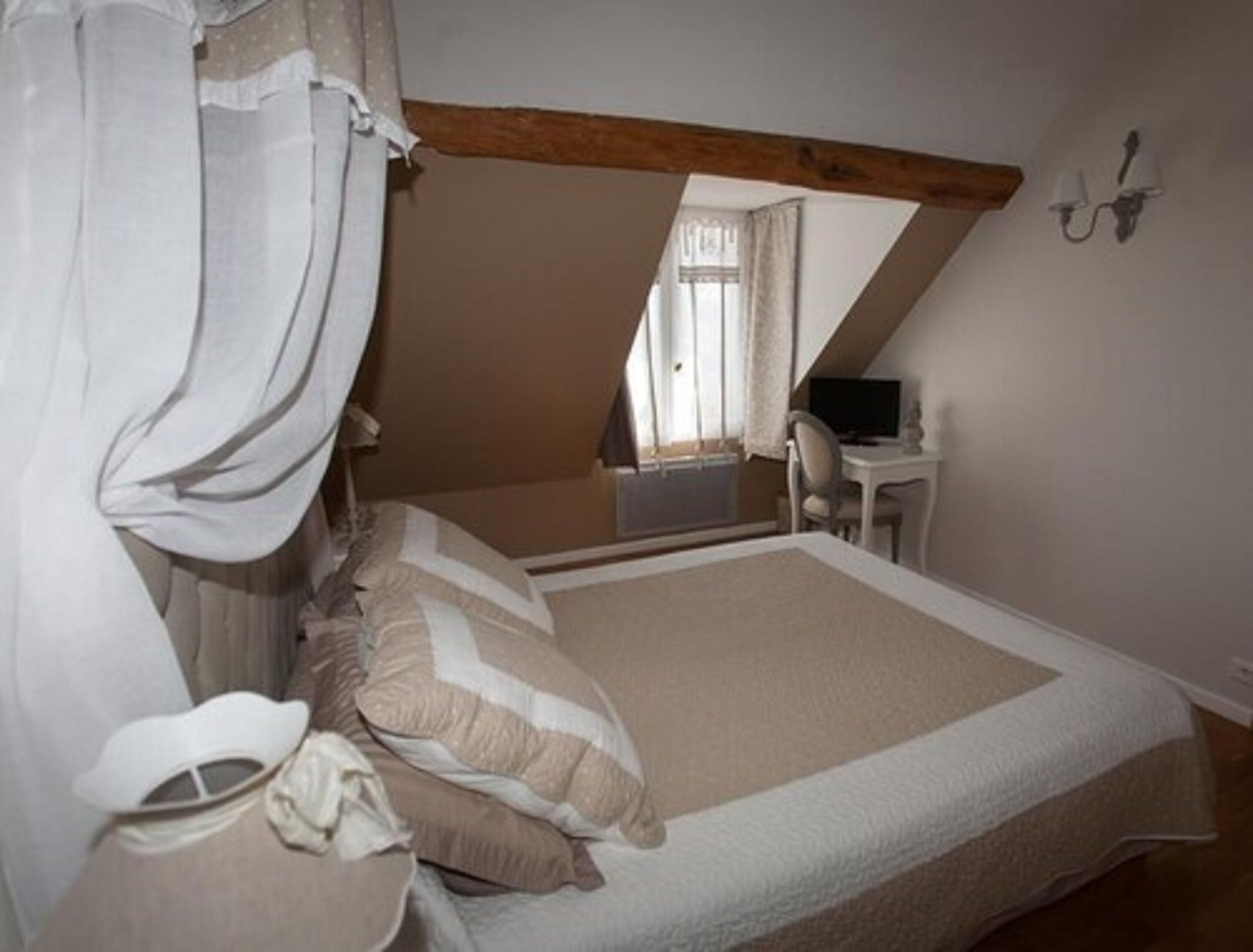 Un dormitorio con techo inclinado, cama con ropa de cama blanca y beige, mesita de noche, una silla y un escritorio con una computadora.