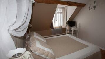 Un dormitorio con techo inclinado, cama con ropa de cama blanca y beige, mesita de noche, una silla y un escritorio con una computadora.
