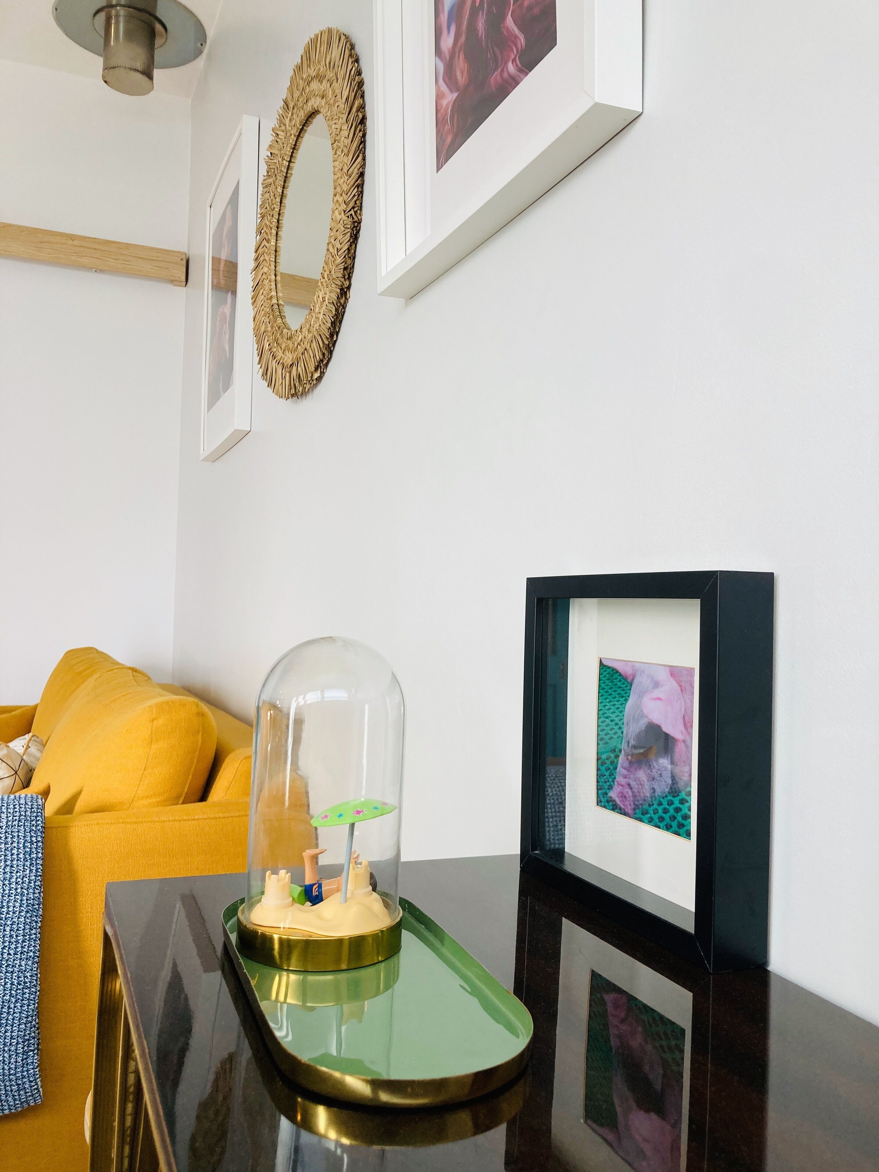 A room with a yellow sofa, a glass dome with a miniature scene, framed pictures on the wall, and a wooden shelf.