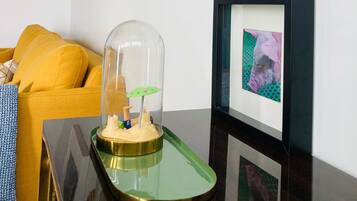 A room with a yellow sofa, a glass dome with a miniature scene, framed pictures on the wall, and a wooden shelf.