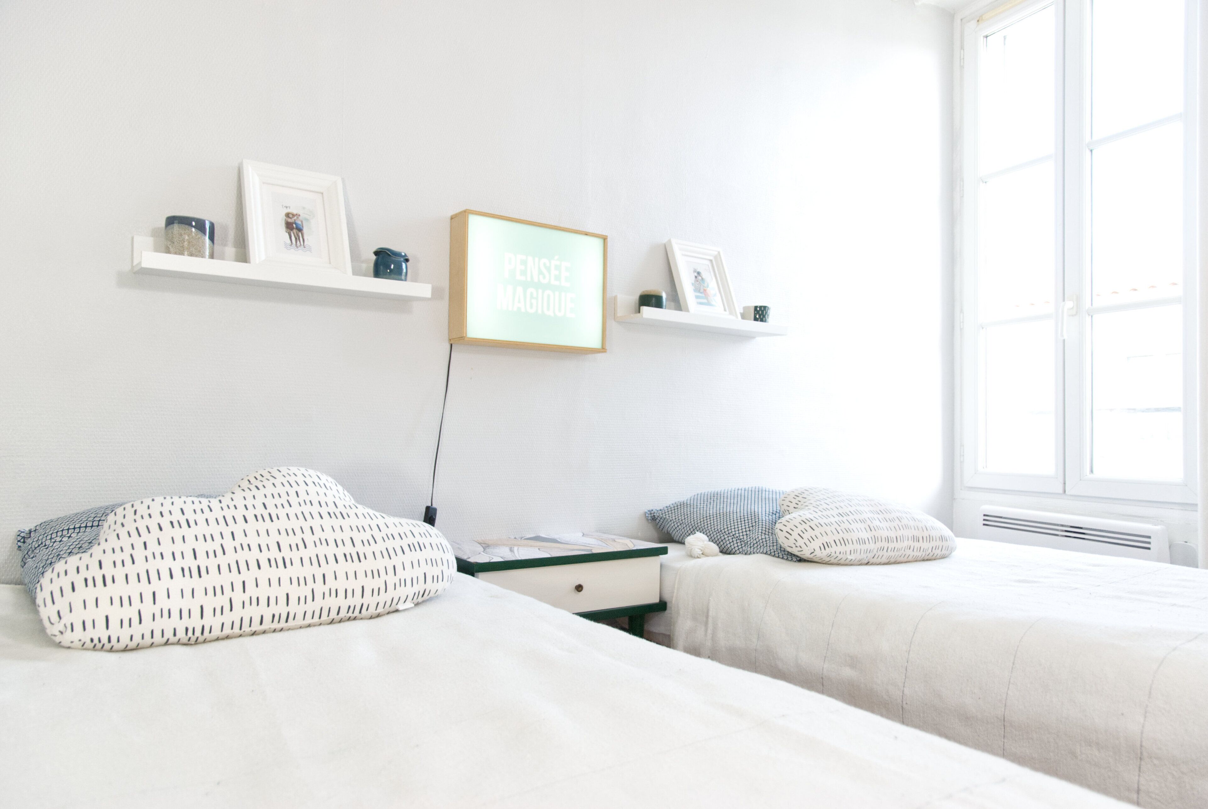 Un dormitorio con dos camas, ropa de cama blanca, una mesita de noche y un estante en la pared con cuadros y un letrero.