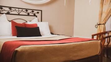 Una cama bien hecha con un edredón rojo, almohadas blancas y una almohada negra. Se ve una silla de madera a la derecha.