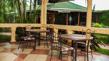 An outdoor patio area with tiled flooring, multiple tables and chairs, and a covered section with a green roof.