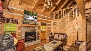 L’intérieur d’une cabane en rondins, avec une cheminée, des canapés en cuir, un téléviseur et un escalier en bois.