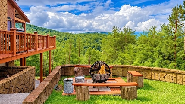 Un patio con un brasero, paredes de piedra y una terraza de madera con vistas a un paisaje boscoso.
