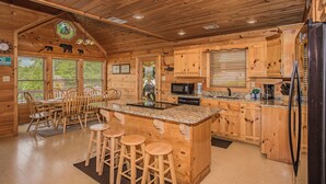 Interior de una cabaña de madera con cocina, que incluye armarios de madera, una isla central con taburetes, una mesa de comedor con sillas y un ventanal con persianas.