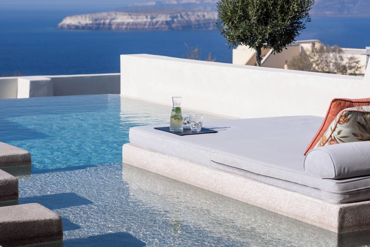 An infinity pool with a clear view of the sea and cliffs, a small table with a glass of water, and a white cushioned bench.