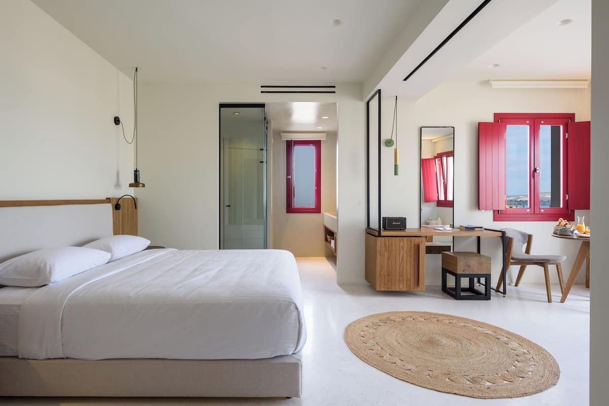 A modern hotel room with a bed, desk, chair, and a view of the sea through a window.