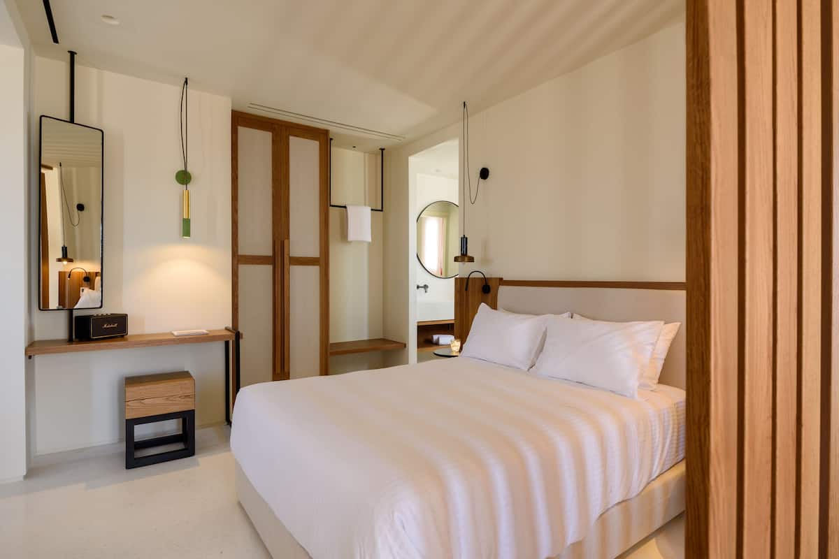 A modern bedroom with a large bed, a bedside table, a mirror, and a small wooden stool.