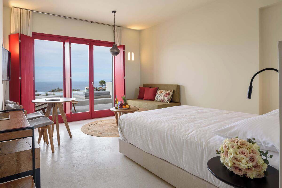 Ein Hotelzimmer mit einem großen Bett, einem Schreibtisch, einem Stuhl, einem kleinen Tisch mit Früchten und Blick auf das Meer durch eine Schiebetür.