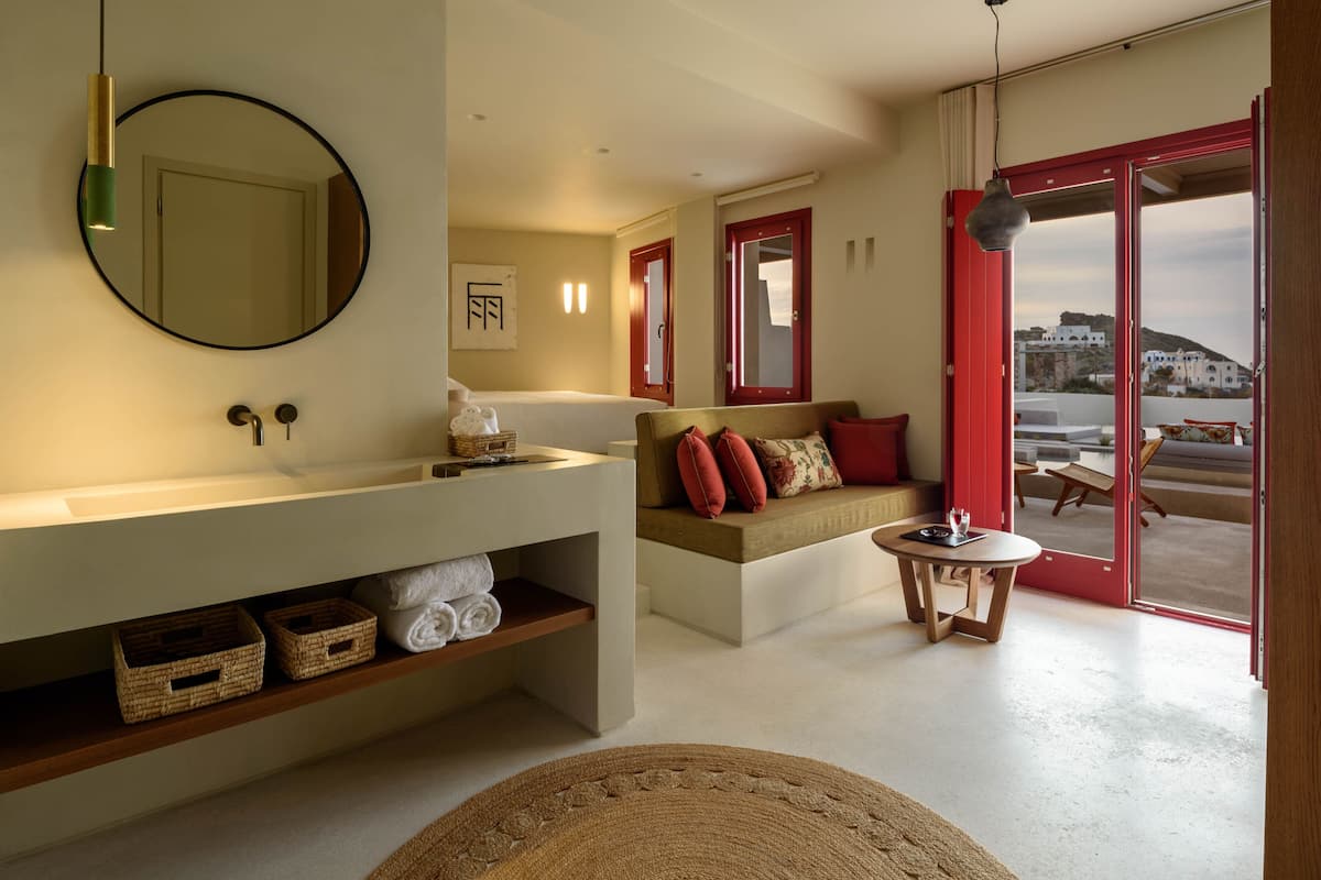 A modern bathroom with a large mirror, a white sink, and a wooden shelf with towels. There is a red-framed door leading to a balcony with a view of buildings.