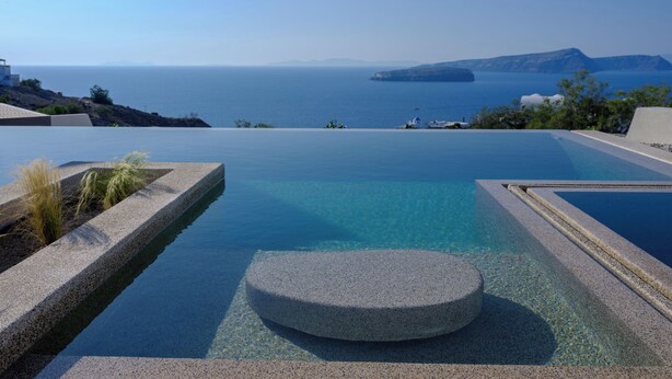 Uma piscina infinita azul cristalina com um elemento circular de pedra no centro, rodeada por uma área ajardinada com plantas.