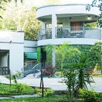 Un edificio moderno de varios niveles con una sección central circular, rodeado de abundante vegetación y un pequeño puente.