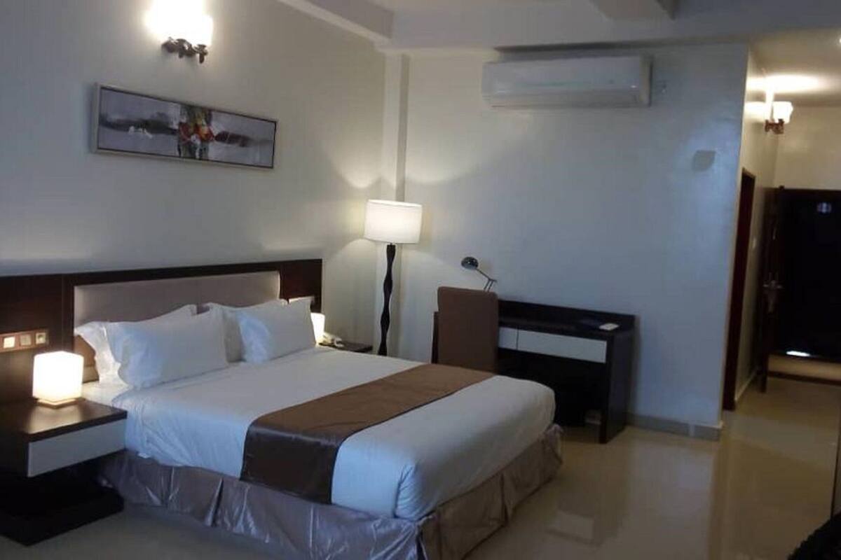 A hotel room with a bed, bedside tables, a desk, a lamp, an air conditioner, and a painting on the wall.