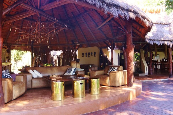 An outdoor lounge area with a thatched roof, leather sofas, and gold-colored stools. - Camp Kwando (Kongola)