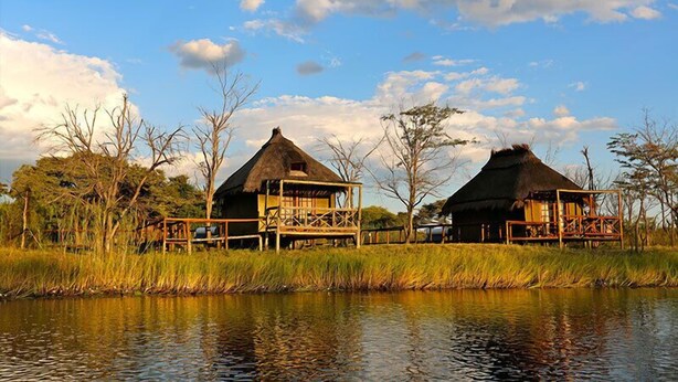 Two thatched-roof structures with wooden decks, situated by a calm body of water, surrounded by tall grasses and sparse trees. - Camp Kwando (Kongola)