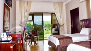 A hotel room with two beds, a desk with a chair, a TV, and a large sliding glass door leading to a balcony with a view of greenery. - Icon Hotel (Chingola)