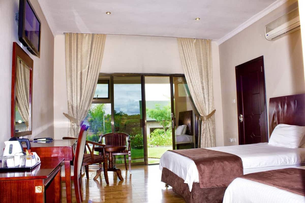 A hotel room with two beds, a desk with a chair, a TV, and a large sliding glass door leading to a balcony with a view of greenery.