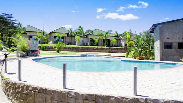 A pool area with a stone border, surrounded by buildings with green roofs and palm trees. - Icon Hotel (Chingola)