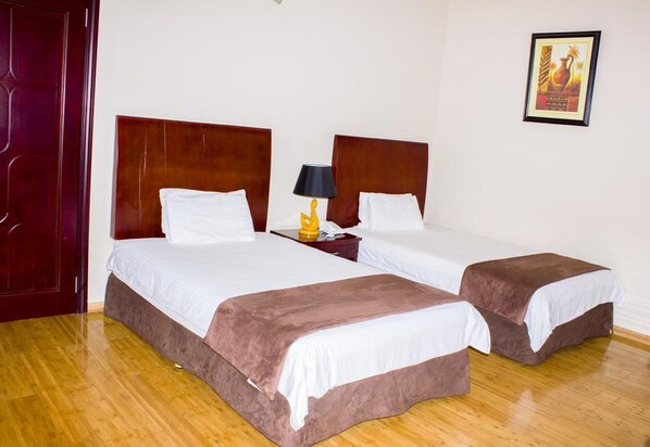 A hotel room with two beds, wooden headboards, white bedding, a nightstand with a lamp, and a framed picture on the wall.