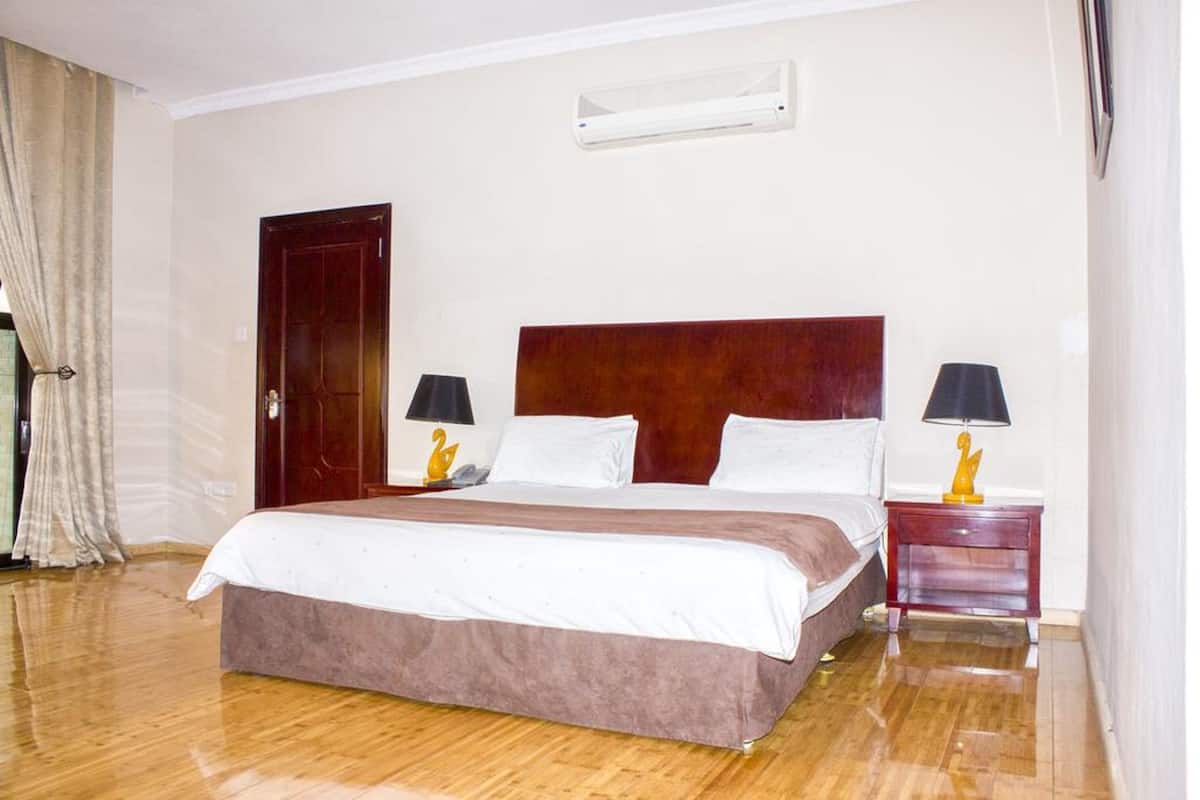 A hotel room with a large bed, two bedside tables with lamps, a wooden headboard, a door, and an air conditioning unit on the wall.