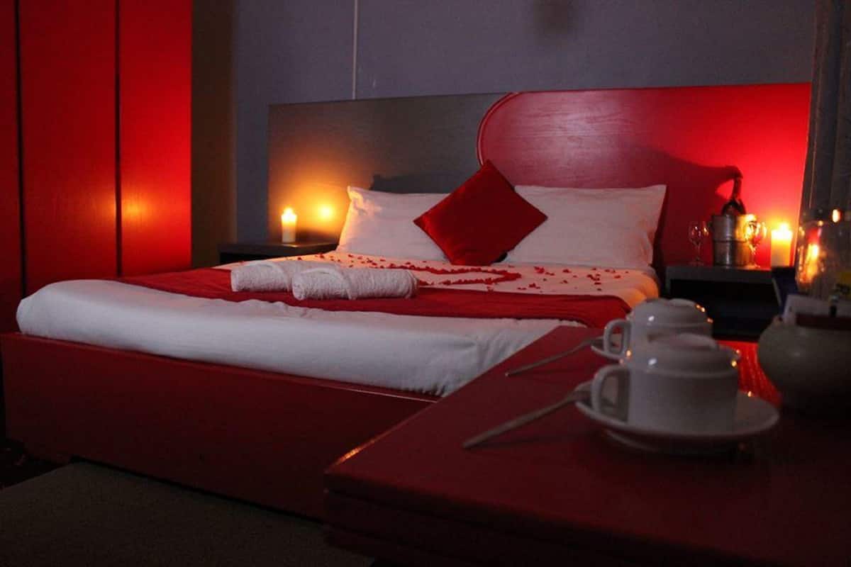 A hotel room with a red bedspread, white linens, a red pillow, and a bedside table with a teapot, cups, and a tray.