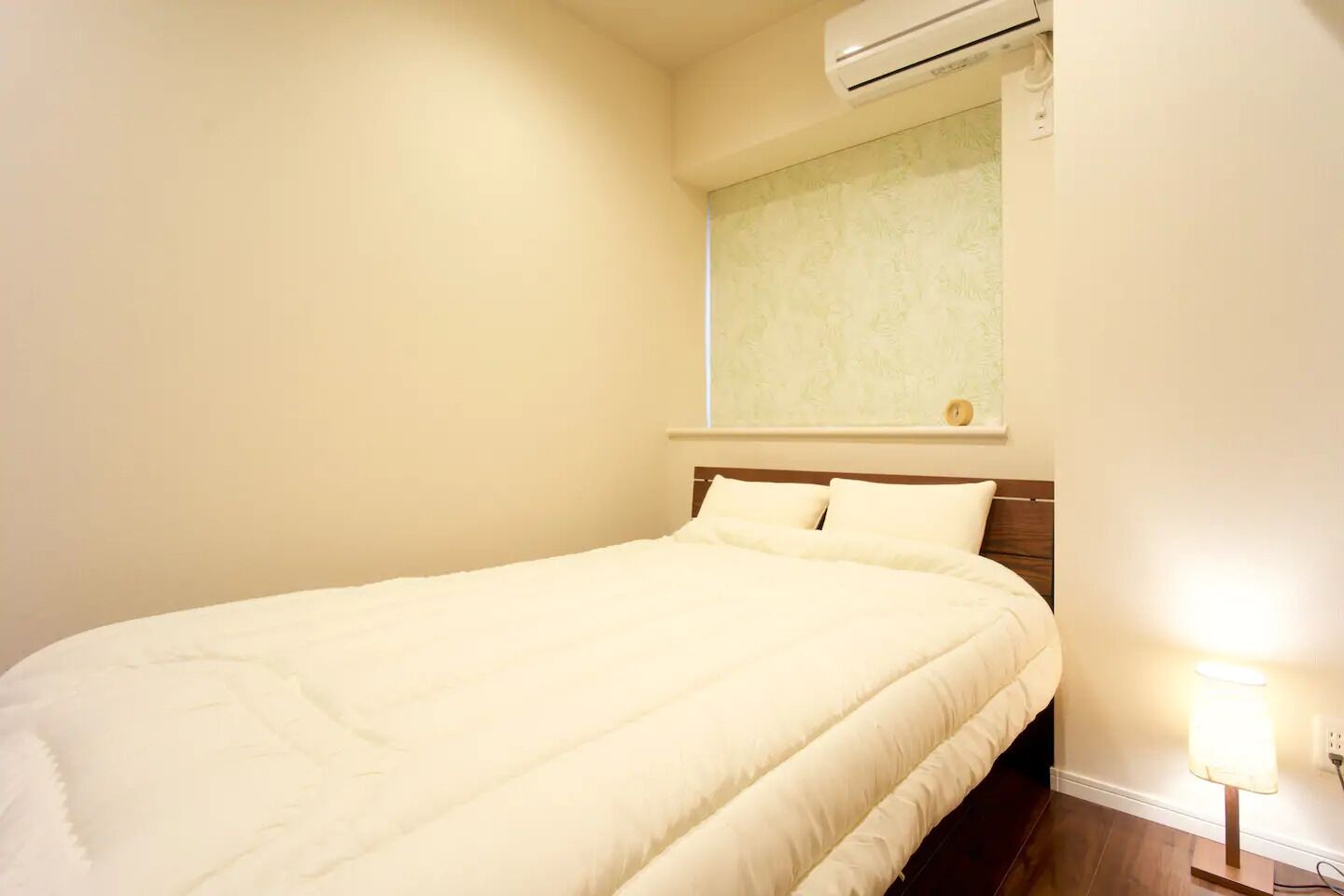A neatly made bed with a white duvet in a room with light-colored walls and a window with a green patterned blind.