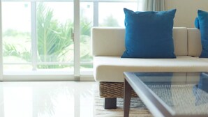 A modern living room with a white sofa, two blue cushions, a glass coffee table, and a view of greenery through large windows. - Longvacation Okinawa 103 (Onna)