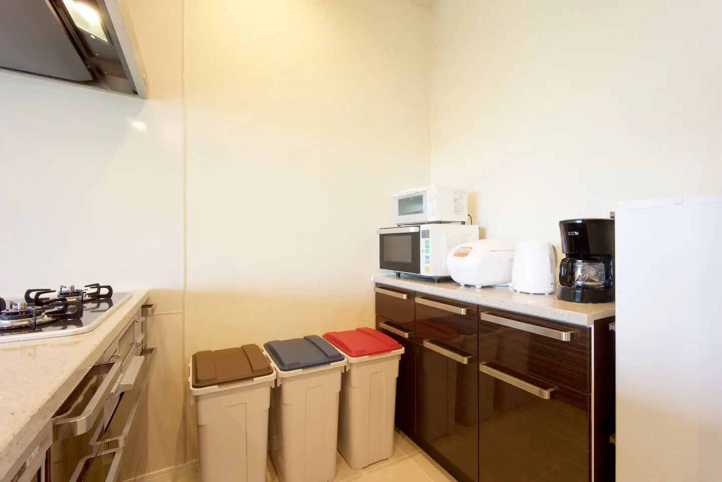 A compact kitchen with a gas stove, microwave, toaster, and coffee maker. There are three trash bins and a small refrigerator.