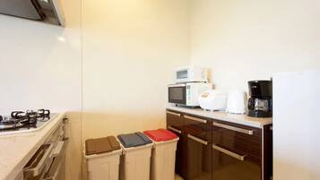 A compact kitchen with a gas stove, microwave, toaster, and coffee maker. There are three trash bins and a small refrigerator.