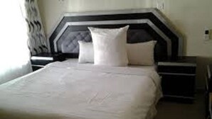 A neatly made bed with a white duvet and pillows, a dark headboard, and a nightstand with a lamp. - Muuka Gaderns lodge (Ndola)